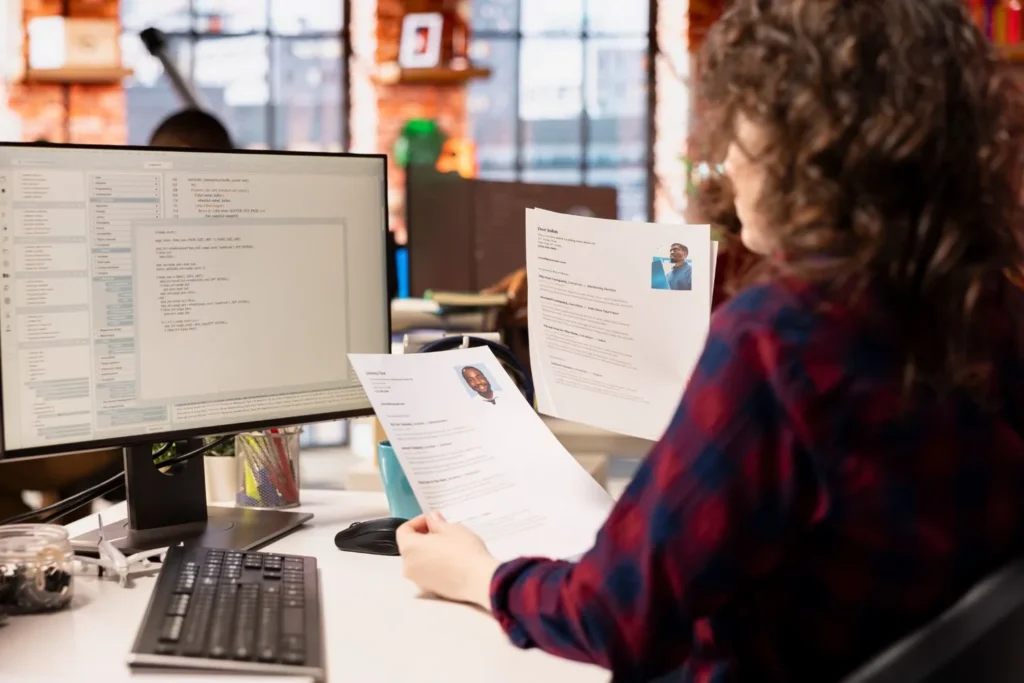 HR professionals reviewing printed resumes while using recruitment software on a desktop computer in a modern office setting.