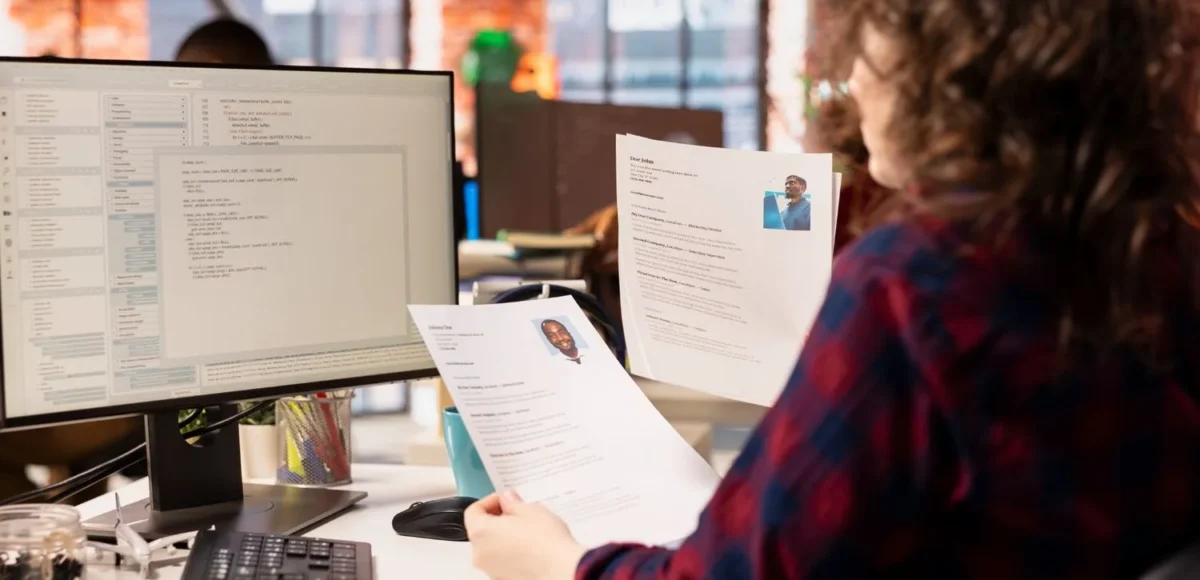 HR professionals reviewing printed resumes while using recruitment software on a desktop computer in a modern office setting.